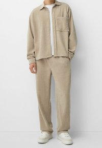 Conjunto de camisa y pantalones de sarga beige con un diseño texturizado y acanalado. Incluye un bolsillo en el pecho y se complementa con zapatillas blancas.