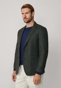 Pánsky blazer z tmavozelenej látky, jednoradová konštrukcia, klopové reverzné límce, predné vrecká, v kombinácii s modrým sveterom s okrúhlym výstrihom a krémovými nohavicami.