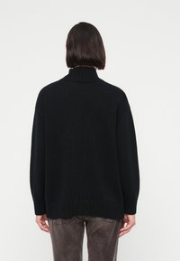 Personne de dos portant un pull à col roulé côtelé noir et un pantalon gris devant un fond blanc uni.
