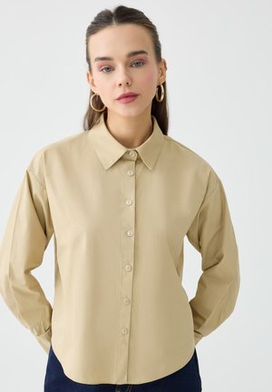 Femme aux longs cheveux bruns portant de grandes boucles d'oreilles en anneau et une chemise beige à boutons, debout avec les mains dans le dos sur fond blanc.