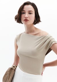 Beige off-shoulder-top med plisserede detaljer og strækbart stof; båret med en struktureret taske. Modellen har kort hår og minimale accessories.