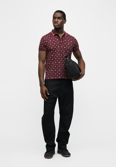 Άνδρας που φοράει μπορντό πόλο με σχέδιο, μαύρο παντελόνι, μαύρα παπούτσια και μαργαριταρένιο κολιέ, κρατώντας μια μαύρη τσάντα μπροστά σε απλό φόντο.