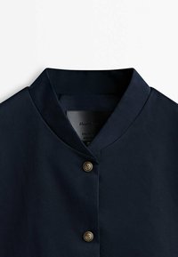 Veste bleu marine avec col mandarin, présentant une texture lisse et deux boutons dorés à l'avant. Design simple et minimaliste.