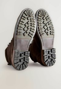 Brązowe zamszowe buty z szorstkimi szarymi gumowymi podeszwami, z teksturowanym bieżnikiem zapewniającym przyczepność oraz solidnym okrągłym noskiem.