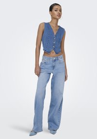 Vrouw draagt een mouwloze gewatteerde spijkervest met knopen, lichtblauwe wijde jeans en blauwe puntige schoenen, staand tegen een effen achtergrond.