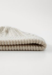 Pehmeast kreemivärvi lõngast kootud beanie, millel on soonikkoes tekstuur ja üle keeratud serv, näidates sujuvat ja ühtlast kootud mustrit.