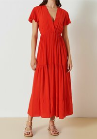 Robe maxi rouge avec un décolleté en V plongeant, des manches courtes flottantes, et un ourlet à volants en étages. Fabriquée en tissu léger, accompagnée de sandales décoratives.