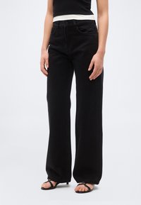 Person trägt schwarze, hochtaillierte Jeans mit weitem Bein, kombiniert mit schwarzen, rückenfreien Sandalen mit Absatz und einem ärmellosen schwarz-weißen Oberteil.