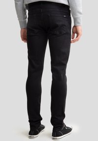 Vīrietis tērpies melnās šaurās džinsās, pelēkā džemperī un melnās sporta kurpēs, stāvot pret vienkāršu gaiši pelēku fonu.