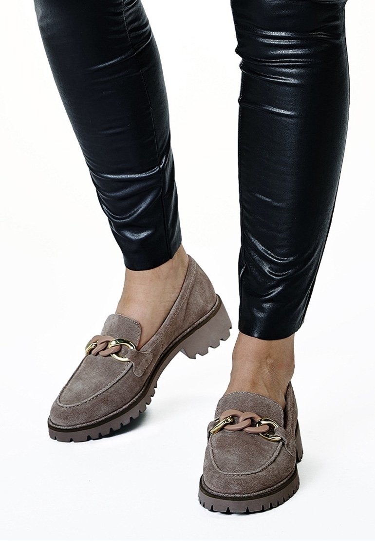 Femme portant des mocassins en suède taupe à semelles épaisses avec un détail chaîne dorée, associés à un pantalon noir aspect cuir.