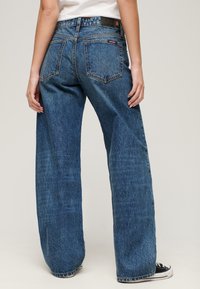 Jeansy denimowe o szerokich nogawkach w ciemnoniebieskim kolorze z lekko wypłowiałą teksturą. Posiadają dwie tylne kieszenie oraz skórzaną metkę z logo na pasie.