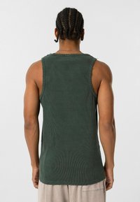 Donkergroen geribbeld tanktop met rauwe randen, mouwloos ontwerp, textuurstof en een getailleerde pasvorm. Achteraanzicht toont details van schouder en arm.