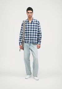 Καρό πουκάμισο σε μπλε και άσπρο, συνδυασμένο με ανοιχτό μπλε τζιν και λευκά αθλητικά παπούτσια. Το μοντέλο κρατάει μια μπεζ τσάντα χεριού.