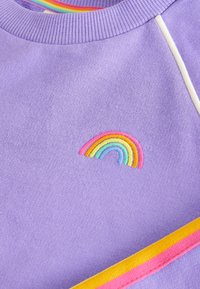 Lila Sweatshirt mit einem kleinen, bestickten Regenbogen in Pastellfarben. Gerippter Halsausschnitt mit kontrastierenden gestreiften Details in Orange, Pink und Blau.