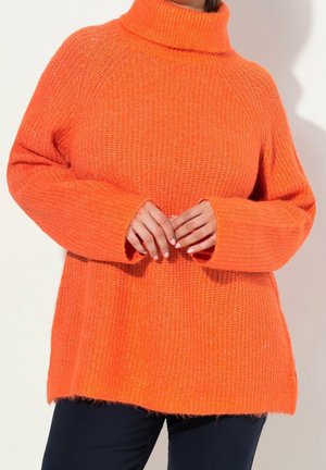 Femme portant un pull à col roulé tricoté épais de couleur orange vif avec des manches longues, associé à un pantalon foncé, les mains doucement jointes.