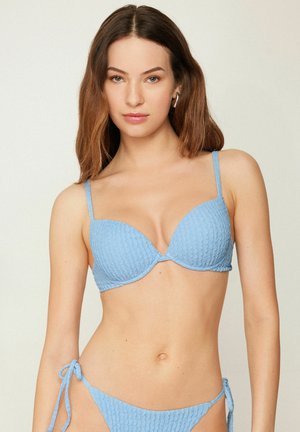 Woman wearing light blue textured bikini with tied sides, standing against a plain neutral background, facing forward.