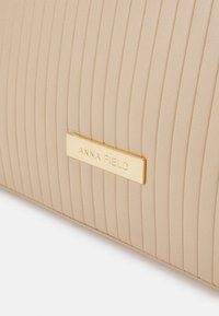 Beige ribbed clutch bagi, jossa on teksturoitu pinta. Varustettu kullalla korostetulla suorakulmaisella logo-laatalla, jossa on teksti "ANNA FIELD."