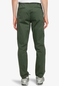 Pantaloni in cotone verdi con vestibilità dritta, dotati di tasche posteriori e una texture liscia. Abbinati a scarpe sportive di colore chiaro.