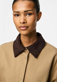 Bež srajca z темnim rjavim žakardnim ovratnikom, ki ima gumbasto zapiranje in teksturirano površino. Tovrstna fotografija prikazuje ovratnik središča.