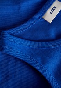 Blue ribbed fabric of a tank top, featuring a smooth neckline and a tag with "JJXX" in a contrasting color. Textured detail is visible.
