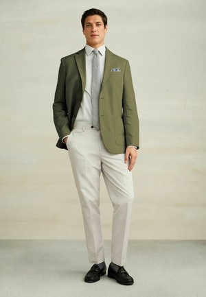 Mann stehend, trägt einen olivgrünen Blazer, ein hell gestreiftes Hemd mit grauer Krawatte, beige Hose und schwarze Loafers vor einem einfarbigen Hintergrund.