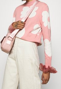 Rosa tröja med stora vita blommönster, ribbad halsringning, tillsammans med off-white, löst sittande byxor och en ljusrosa axelväska.