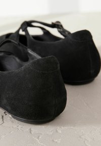 Zwarte suède ballerina's met een ronde neus en achterkant, voorzien van een enkelbandje en een zachte textuur. De zool is stijlvol en plat.