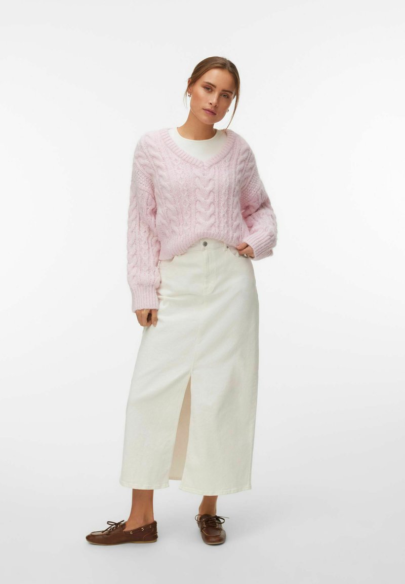 Femme portant un pull en maille torsadée rose clair, une jupe midi blanche taille haute avec une fente frontale, et des chaussures marron à lacets, debout avec une main dans la poche.