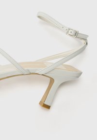 Valkoinen remmillä varustettu korkokenkä, jossa on kapea korko ja beige sisäpohja. Säädettävä nilkkaremmi, jossa on hopeanvärinen solki. Silkkinen pintakäsittely.