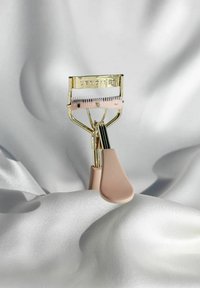 Curling des cils en métal avec une finition dorée et une prise en caoutchouc rose, équipé d'un coussinet en silicone blanc et de dents fines pour courber les cils.