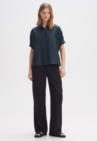 Camicetta verde scuro a maniche corte con bottoni, dal taglio comodo, abbinata a pantaloni larghi neri e sandali neri a scivolo. Design semplice.