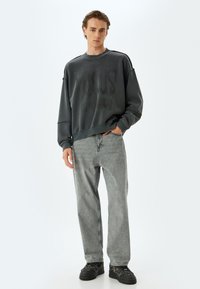 Donkergrijze oversized sweatshirt met een textuurafwerking, voorzien van een logo-print aan de voorkant, gecombineerd met lichtgrijze rechte jeans en zwarte sneakers.