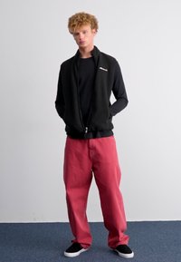 Melnas vilnas veste ar rāvējslēdzēju virs melnas garām piedurknēm krekla, apvienota ar brīvām sarkanām biksēm un melniem slip-on apaviem. Vienkāršs dizains un ikdienišķa piegriezums.