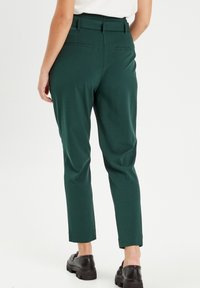 Personne portant un pantalon tapered vert foncé et des mocassins noirs épais, vu de dos sur un fond blanc.