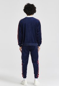 Marinblå velour joggingsdräkt med vit krage, försedda med röd och vita sidostråkar på både jackan och byxorna. Elastiska manschetter och midjeband.