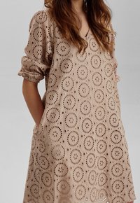 Beige ögla klänning med cirkulära utskärningar, avslappnad passform, korta puffärmar, V-halsringning och sidofickor. Slät textur och andningsdesign.