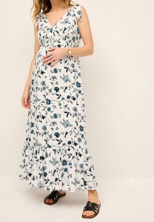 Schwangere Frau trägt weißes florales ärmelloses Maxikleid und schwarze Sandalen, hält sanft mit einer Hand den Bauch.