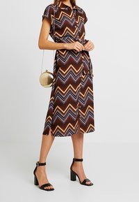 Robe à motifs en chevrons bruns, à manches courtes, avec boutonnière à l'avant et ceinture à la taille. Associée à des sandales noires à talons carrés et à un sac rond doré.