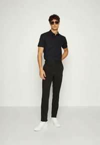 Black short-sleeve button-up shirt, slim-fit black trousers, and white sneakers. Smooth fabric and modern cut. Stylish sunglasses complete the look.