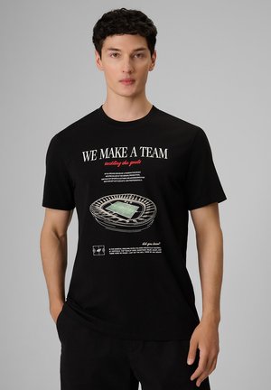 Młody mężczyzna w czarnej koszulce z grafiką stadionu piłkarskiego i napisem "We make a team tackling the goals," stoi na szarym tle.