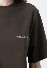 Donkerbruine katoenen t-shirt met korte mouwen en een ronde halslijn, met daarop witte cursieve tekst "Ik blijf laat werken" op de borst.