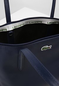 Navy blue tote bag with textured fabric, Lacoste logo, and contrasting internal label. Features dual sturdy handles and a zip closure.