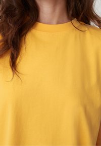 Kobieta ubrana w gładką żółtą koszulkę z okrągłym dekoltem, z długimi brązowymi falującymi włosami częściowo widocznymi po lewej stronie.