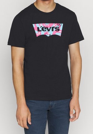 Mężczyzna ubrany w czarną koszulkę Levi's z różowo-niebieskim wzorem flamingów wewnątrz logo marki, w połączeniu z niebieskimi dżinsami.