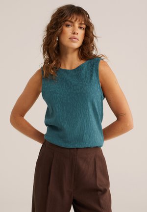 Vrouw met golvend bruin haar, gekleed in een mouwloos teal patroon top en een hooggetailleerde bruine broek, staand met haar handen achter haar rug.