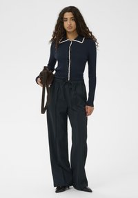 Vrouw met krullend haar die een marineblauwe geribde top met crème rand, losse zwarte broek, zwarte puntige schoenen draagt en een bruine ronde tas vasthoudt.