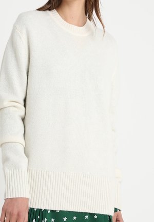 Femme portant un pull en maille blanc avec des poignets et un ourlet côtelés, sur une jupe verte ornée de motifs d'étoiles blanches.