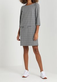 Vestido a cuadros grises con mangas de tres cuartos, que presenta bolsillos con cremallera. Combinado con zapatillas blancas para un look casual.