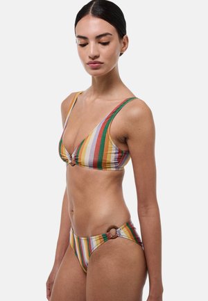 Femme portant un bikini rayé vertical multicolore avec un détail anneau sur la partie basse, debout devant un fond blanc uni.