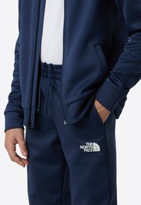 Navy blue zip-up jacket and matching sweatpants made of smooth fabric, featuring an elastic waistband and white logo detail on the pants.